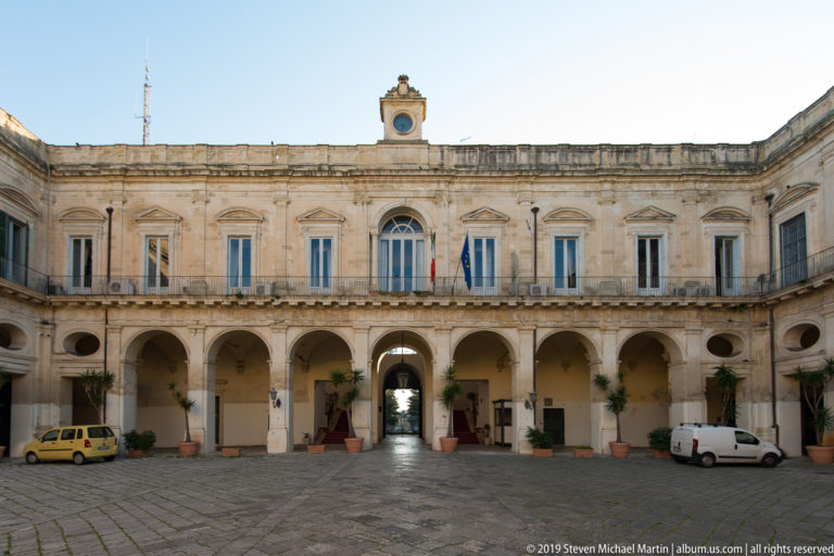 Stone-paved Palazzo della Provincia courtyard with ground-floor arcades, upper windows, flags, potted plants and parked vehicles.