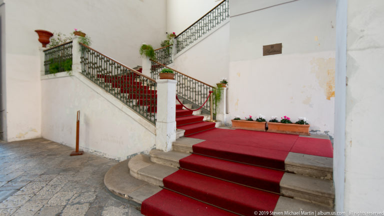 Red-carpeted stone staircase with black wrought-iron railing, potted flowers and pale walls inside a historic civic building.