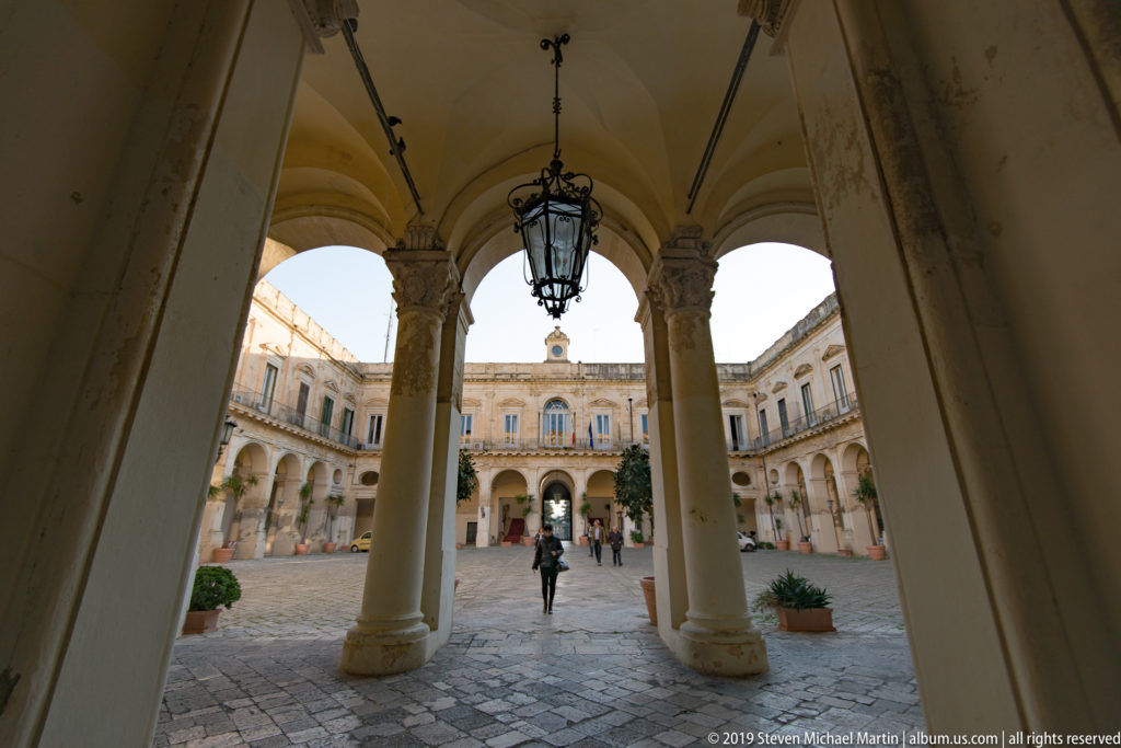 Palazzo della Provincia courtyard seen through a dark arcade, framed by columns and arches with a hanging lantern overhead.