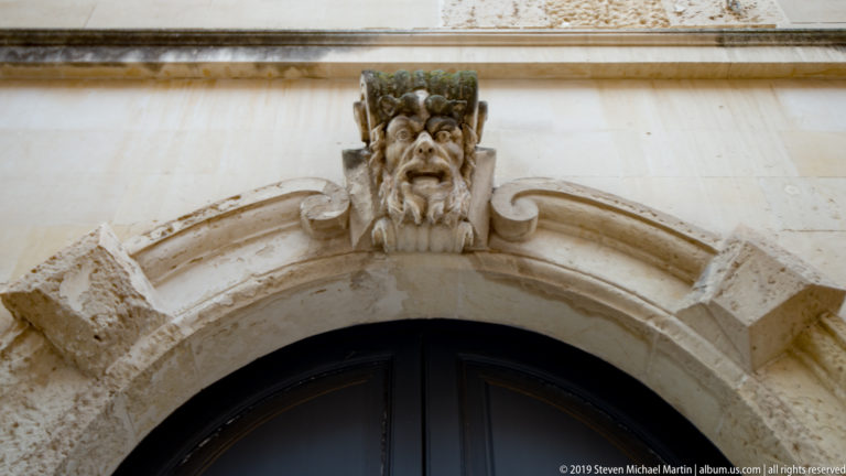 Carved grotesque face with open mouth and leafy crown centered above a rounded stone doorway.