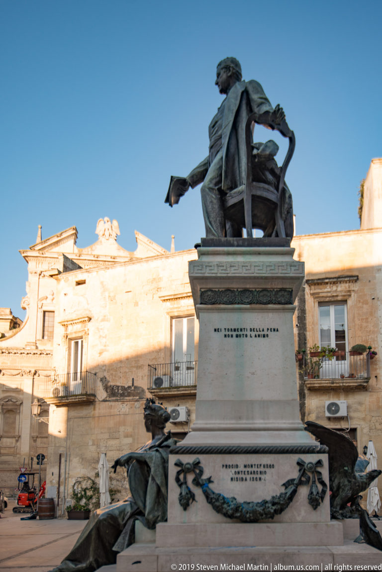 Bronze seated figure atop a tall inscribed stone pedestal with wreath ornament, set against historic Lecce buildings.