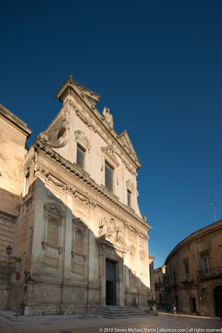 Pale Baroque church façade with pilasters, niches and layered cornices beside a small Lecce square under deep blue sky.