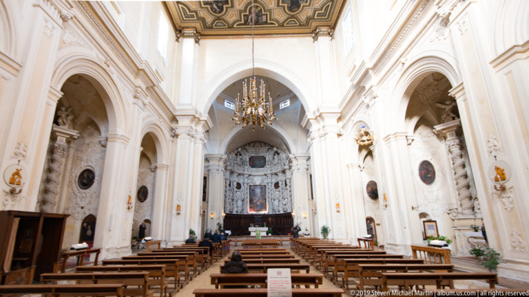 Bright Baroque church nave with wooden pews, side arches, oval paintings, chandeliers and an ornate high altar.