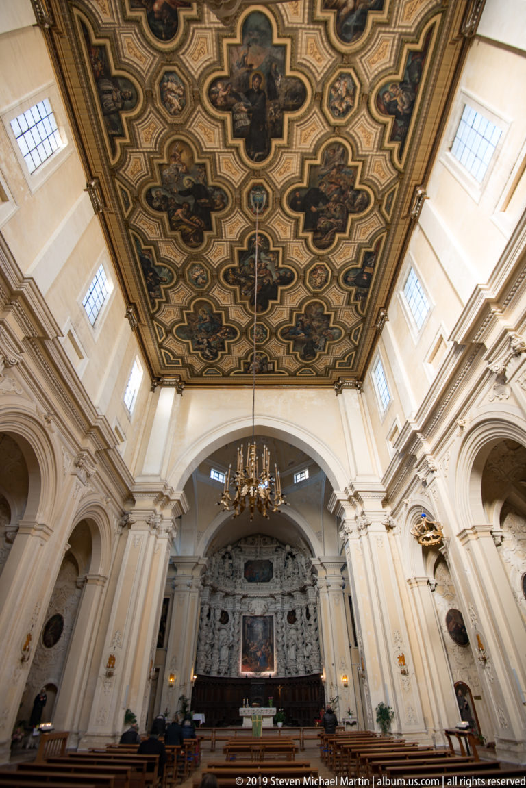 Dark green and gold patterned coffered ceiling extending above a bright church nave and chandelier.