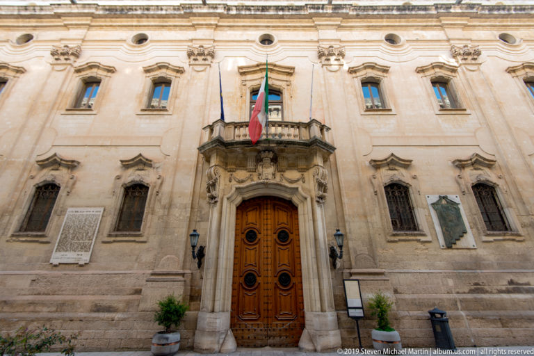 Formal pale-stone civic façade with carved central doorway, balcony, Italian and European flags, and symmetrical windows.