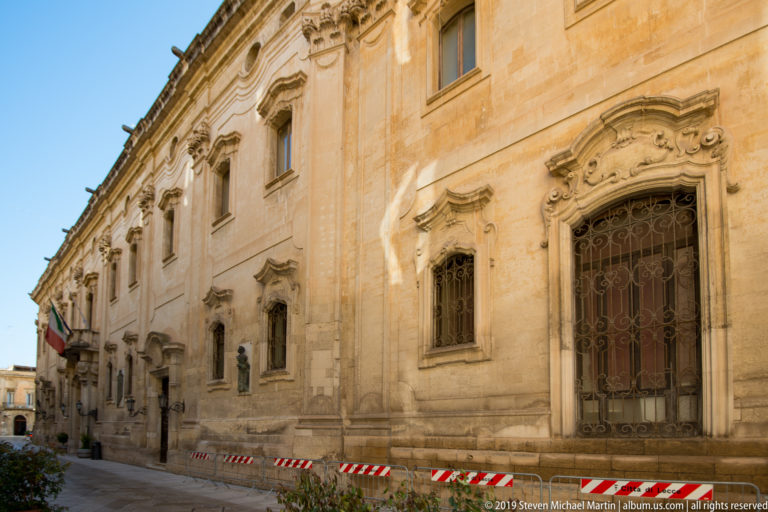 Long pale-stone historic façade with ornate window surrounds, iron grilles, pilasters and temporary barriers along the street.
