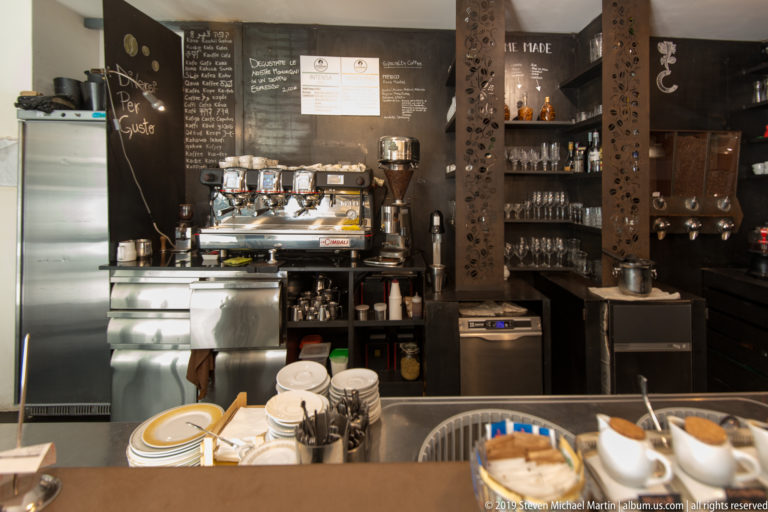 Compact café bar with espresso machine, stacked cups, glassware shelves and handwritten chalkboard menus.