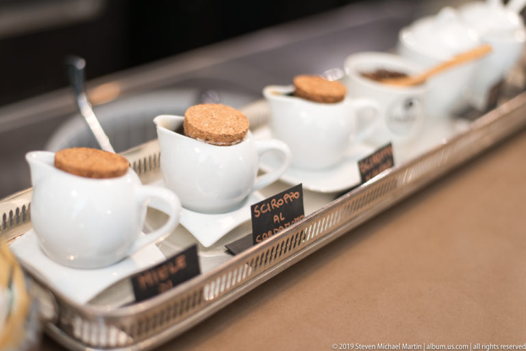 Small white ceramic pots with cork lids arranged on a café tray beside handwritten flavor labels.