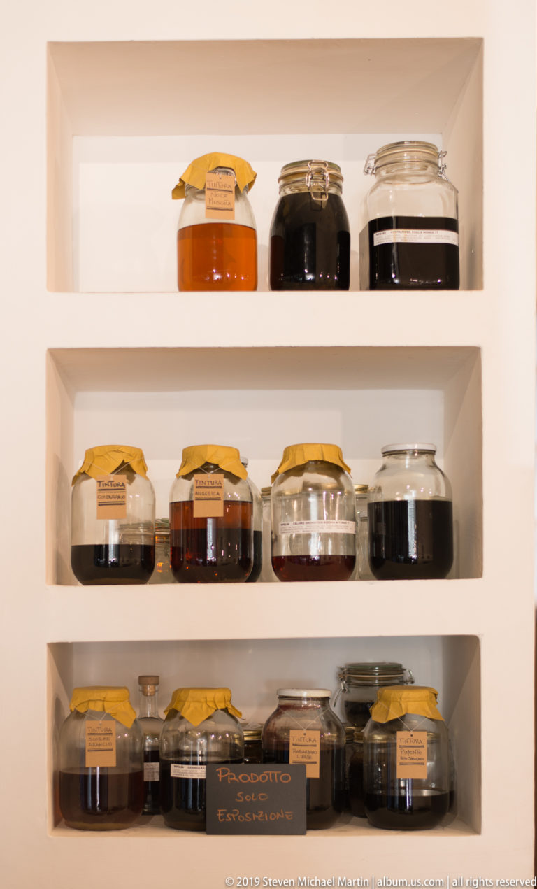 Rows of glass jars containing amber and dark preserves arranged on three recessed white shelves with small labels.