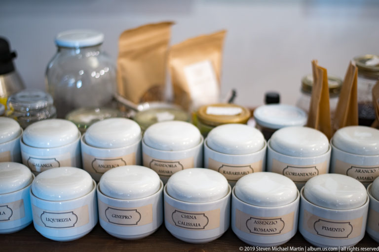 Rows of small white labeled jars for herbs and spices, with glass bottles and paper packets blurred behind them.