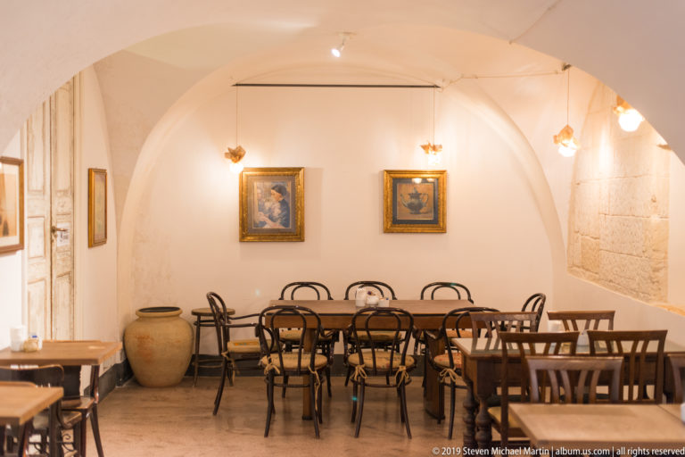 Vaulted white café dining room with wooden tables, bentwood chairs, framed paintings and a large ceramic jar.