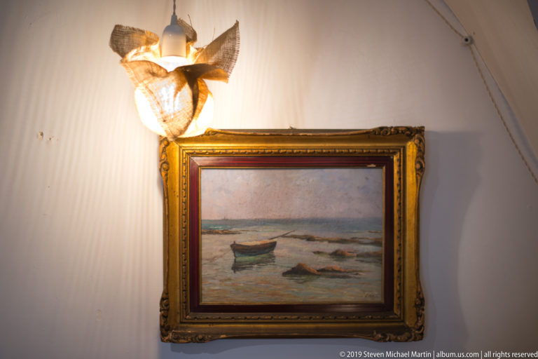 Small framed seascape with boats on water, hung beneath a decorative wall light on a pale interior wall.