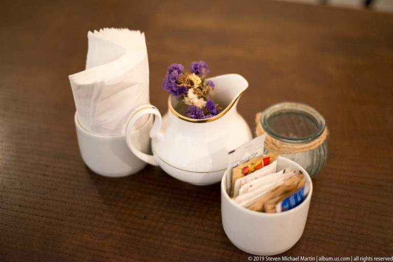 White ceramic café vessels with folded napkins, sweetener packets and purple dried flowers arranged on a wooden table.