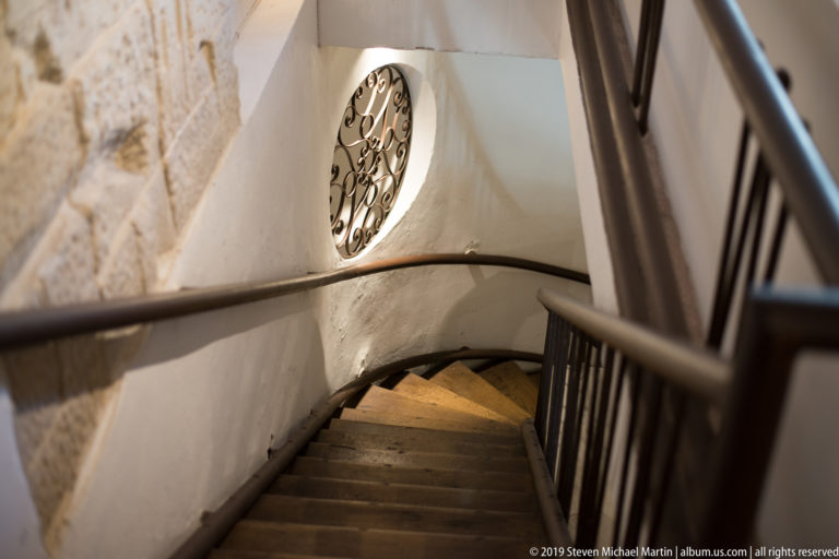 Narrow descending stone staircase with dark handrail, white walls, exposed masonry and a circular decorative iron grille.
