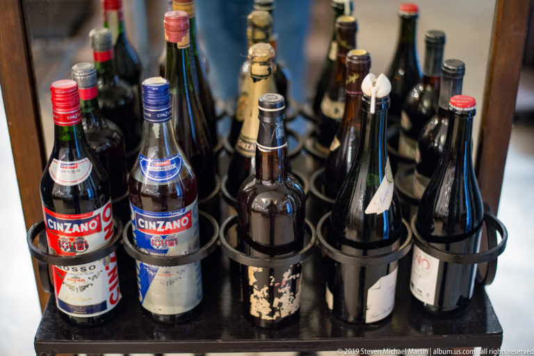 Assorted dark glass beverage bottles in a metal rack, with red- and blue-labeled Cinzano bottles at the front.