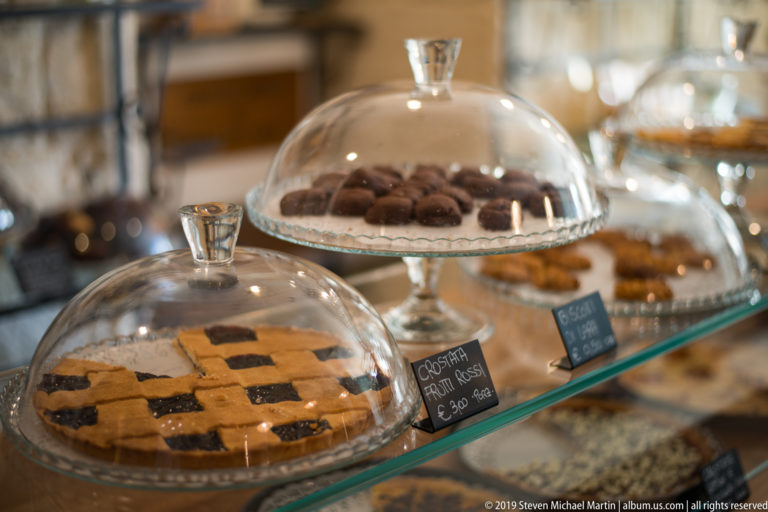 Lattice tart and chocolate pastries displayed beneath clear glass domes on a café counter with small handwritten signs.