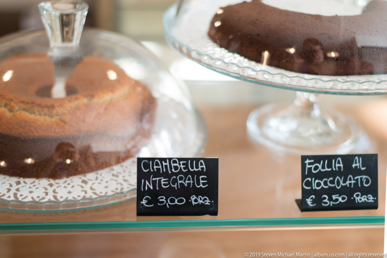 Pale ring cake and chocolate-covered ring cake displayed on glass stands behind handwritten café price signs.