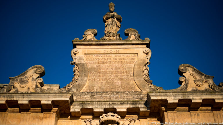 Upper section of Porta Napoli with Latin inscription, sculpted cresting and a standing figure silhouetted against deep blue sky.
