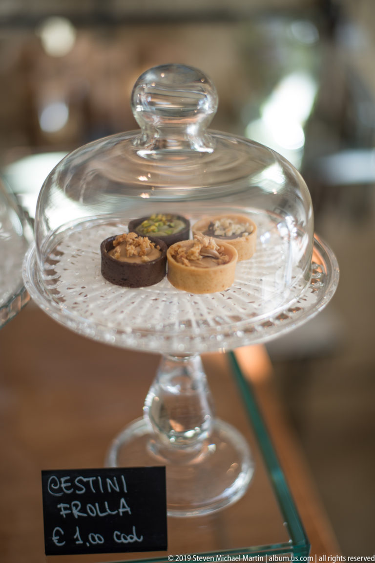 Three small rolled pastries on a patterned glass stand beneath a clear glass cloche, with a handwritten price card below.