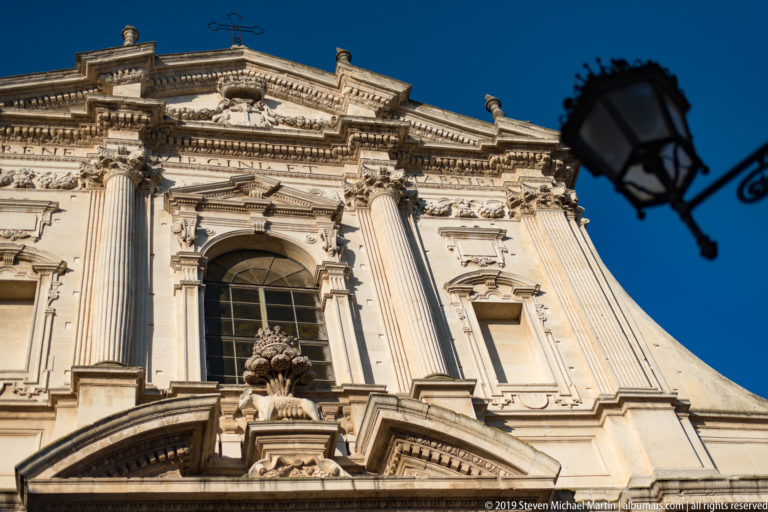 Upward view of ornate pale-stone church façade with fluted pilasters, central arched window, carved ornament and black street lamp.