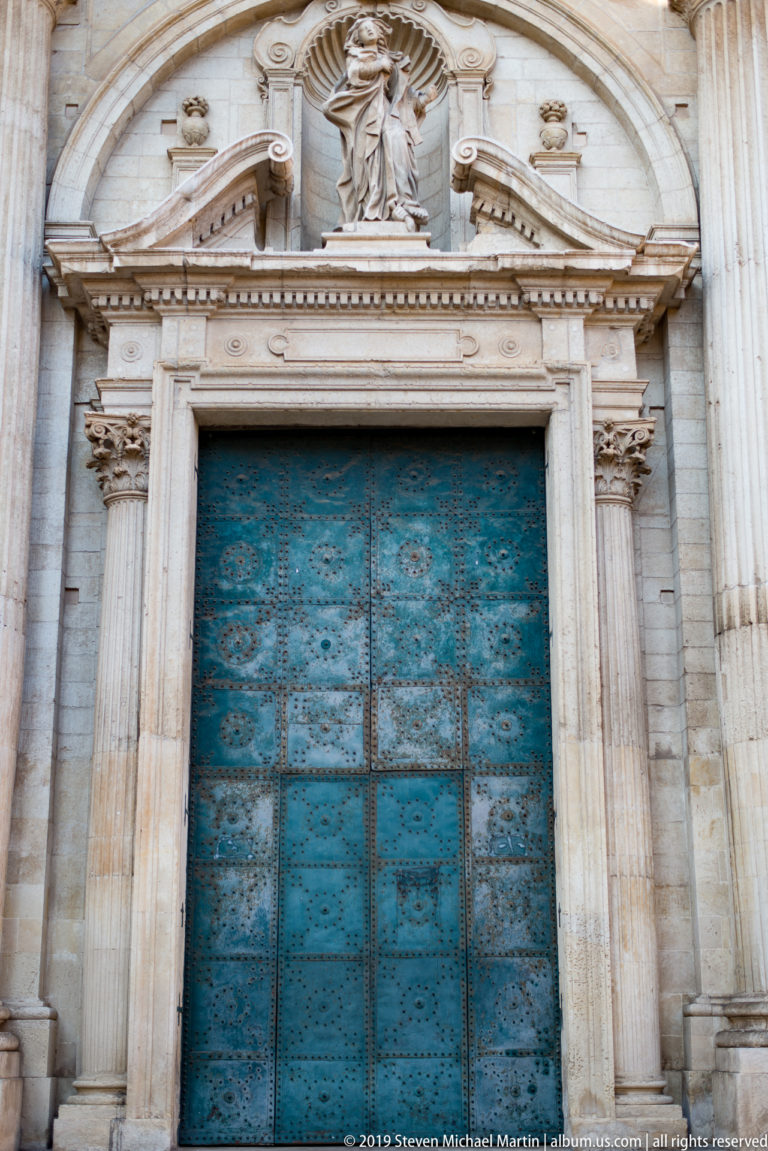 Blue-green metal cathedral doors framed by fluted stone columns, with a sculpted religious figure above the portal.