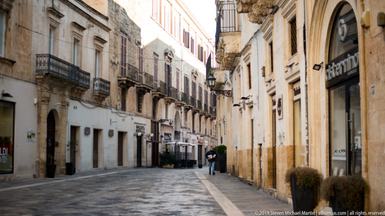 Stone-paved Lecce street lined with pale historic buildings, iron balconies, shopfronts and a few distant pedestrians.