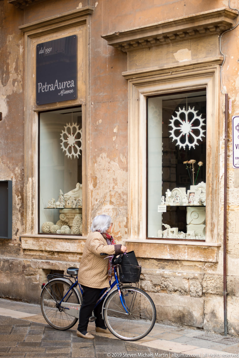 Petra Aurea shop windows displaying pale ceramics as a cyclist passes along the weathered stone façade.