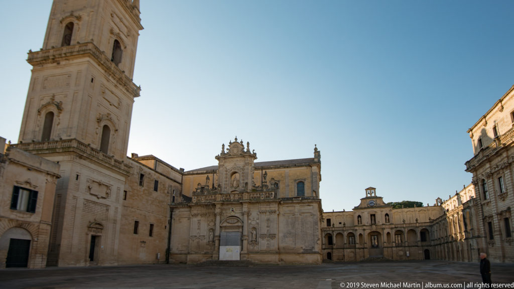 Wide view of Piazza del Duomo with Lecce Cathedral complex, tall bell tower, ornate church façade and arcaded surrounding buildings.