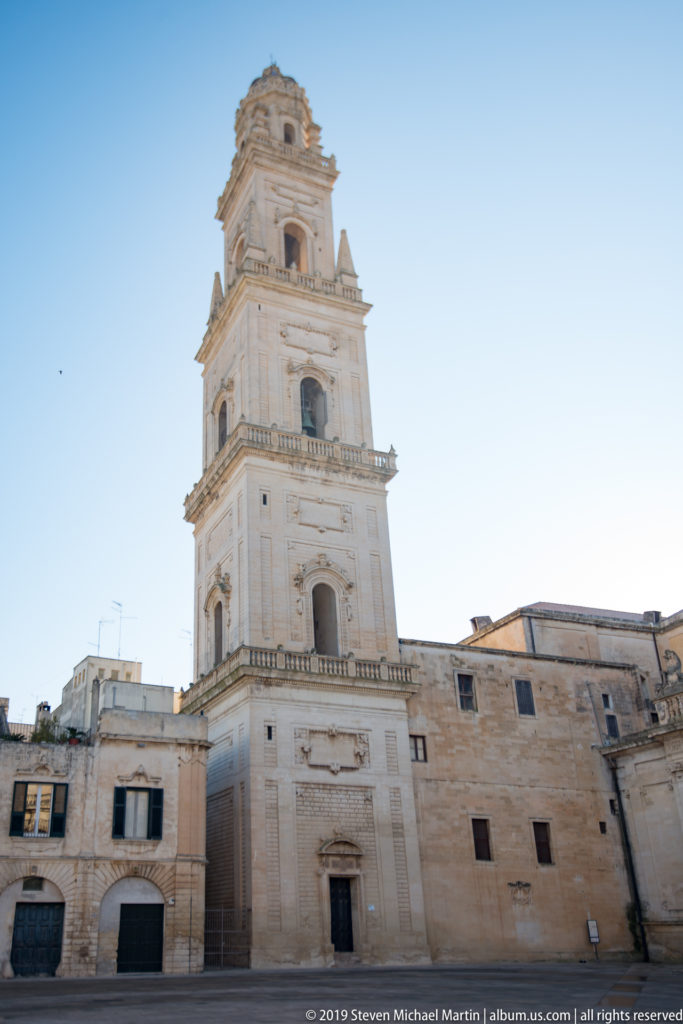 Lecce Cathedral bell tower rising in several pale-stone stages with arched openings above surrounding buildings.