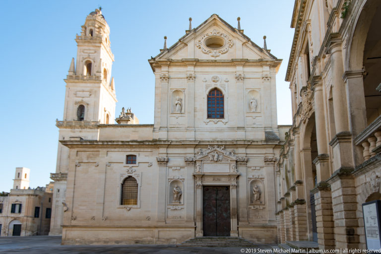Pale limestone church façade with sculpted doorway, upper niches and pediment, beside a square bell tower under blue sky.