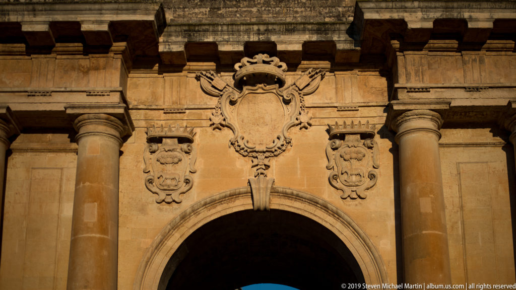 Crowned central shield and smaller carved coats of arms above Porta Napoli’s arch, framed by stone columns.