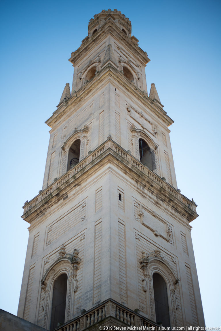 Low-angle detail of Lecce Cathedral bell tower with arched openings, pilasters, balustrades and projecting cornices.