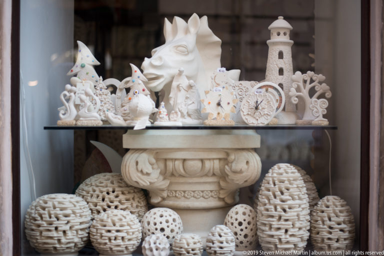 Shop window filled with white decorative ceramics, including pierced egg forms, a lighthouse, clock and stylized horse head.