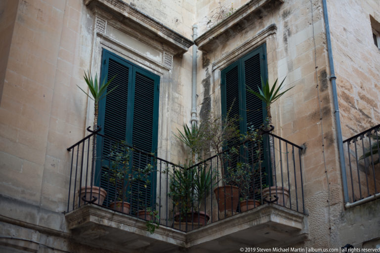 Small corner balcony with dark green shutters, iron railing and potted palms against warm limestone walls.