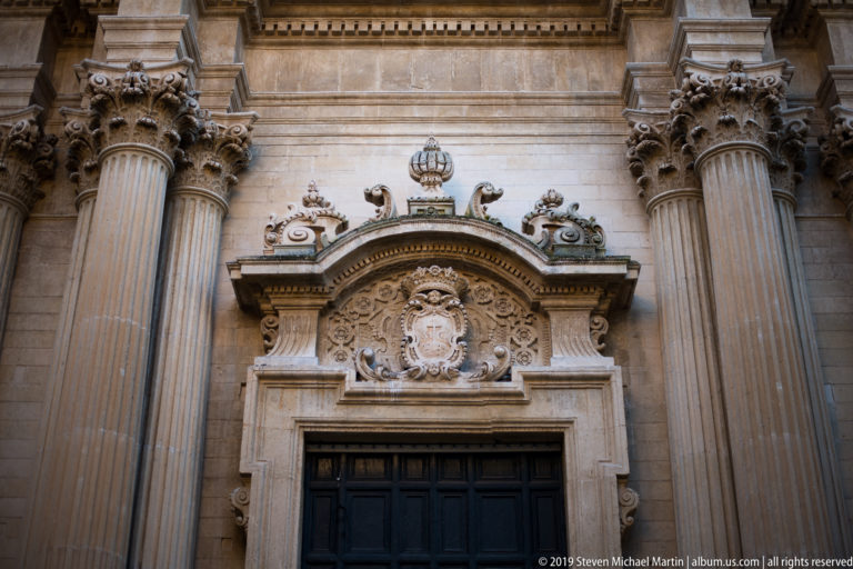 Dark doorway beneath an elaborate crowned heraldic cartouche, framed by giant fluted columns and sculpted Baroque ornament.