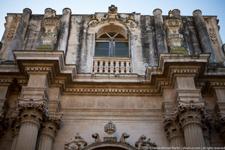Upper limestone façade with central balconied window, paired columns, carved capitals and weathered Baroque ornament.
