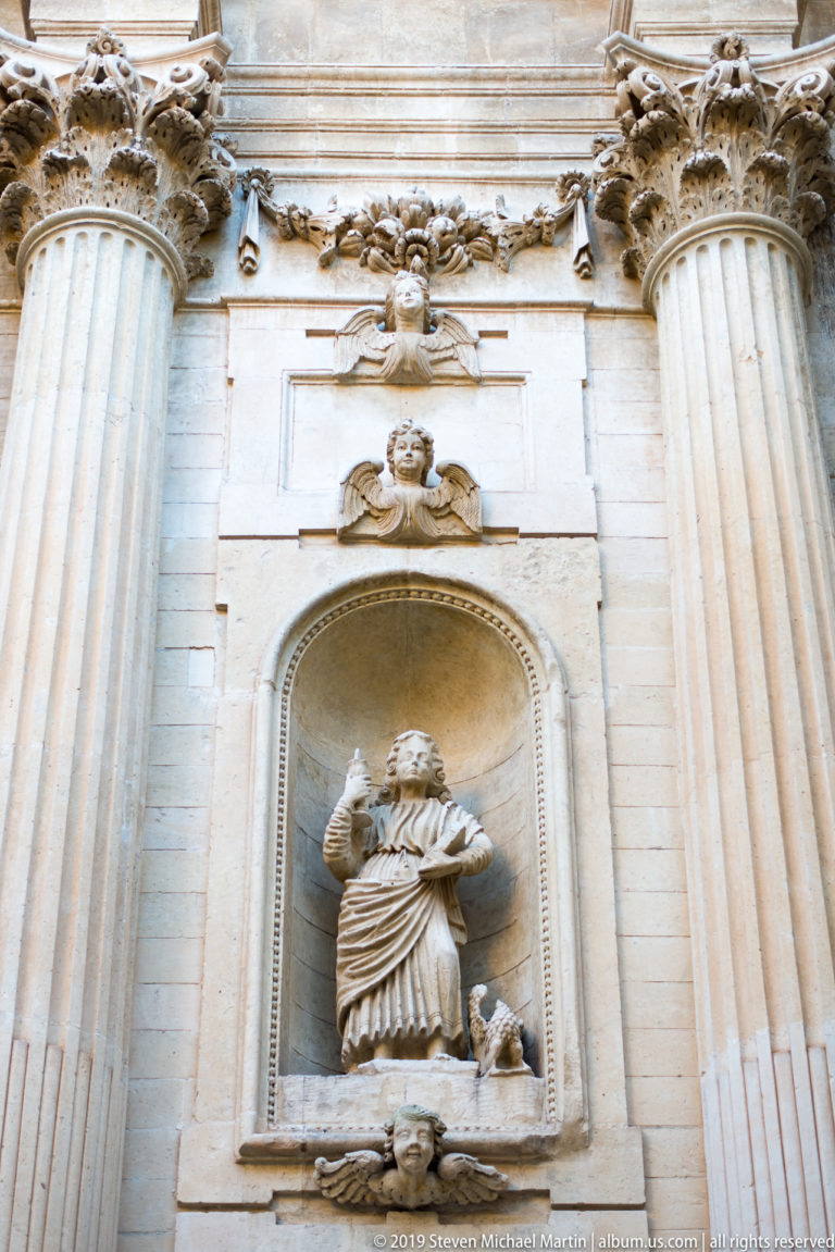 Robed stone religious figure standing in an arched niche between fluted columns, with carved garland above.