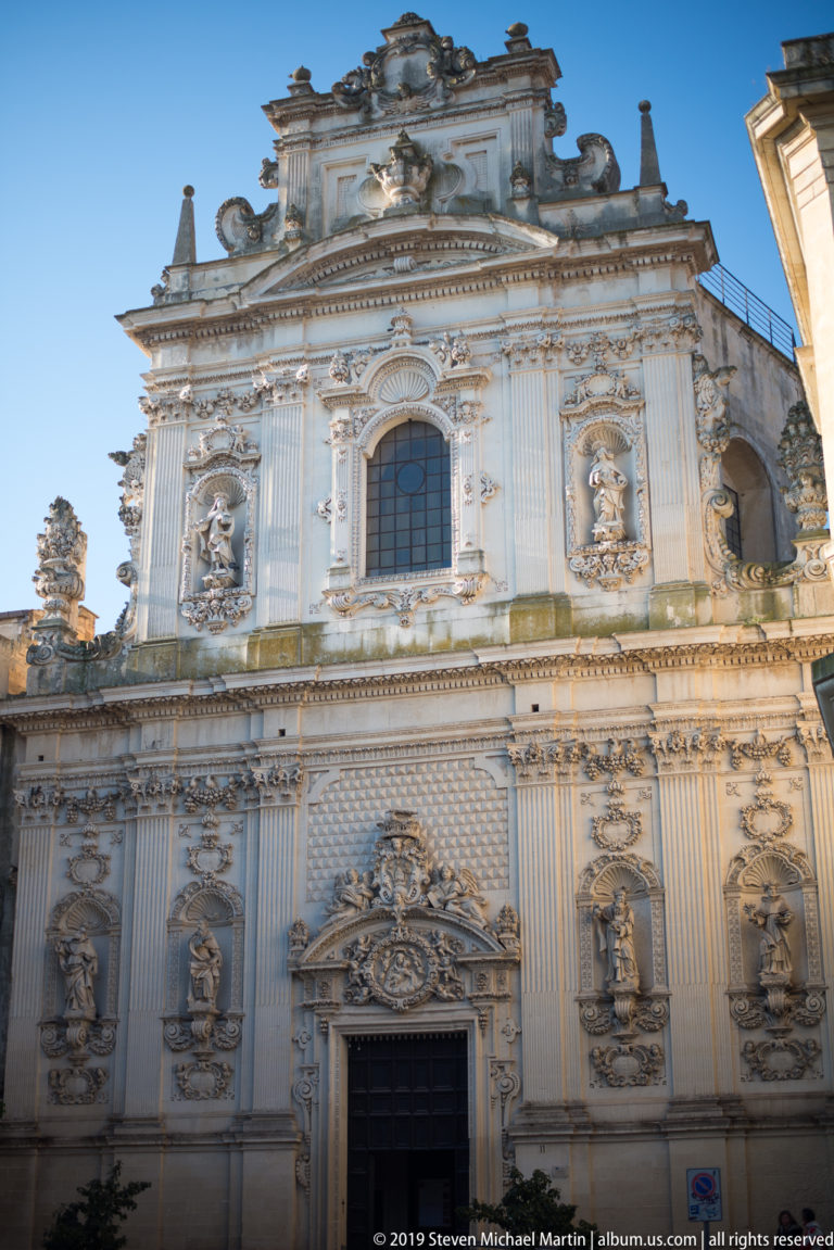 Ornate pale-stone church façade with statues in niches, fluted pilasters, carved portal and elaborate upper pediment.
