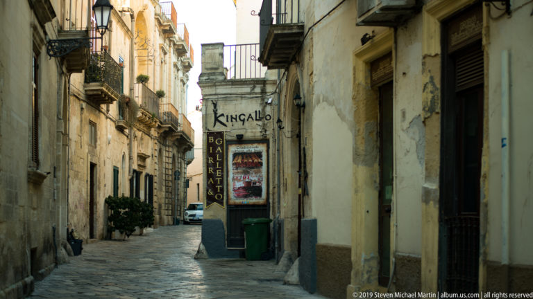 Narrow paved lane between weathered stone buildings with balconies, shutters, signs and a small business entrance.