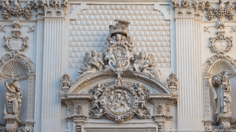 Highly ornate limestone façade with standing saints, fluted pilasters, shell niches, heraldic ornament and central religious relief.