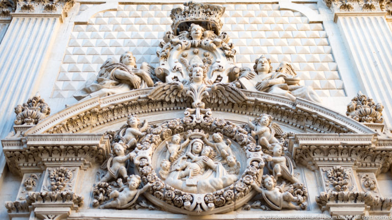 Circular religious relief surrounded by angels and sculpted figures beneath a triangular pediment and elaborate upper cartouche.