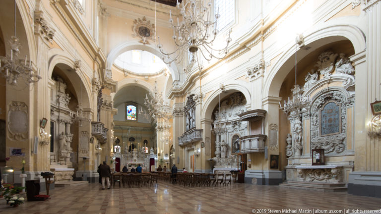 Bright Baroque church nave with chandelier, ornate side altars, arches, paintings and visitors near the distant sanctuary.