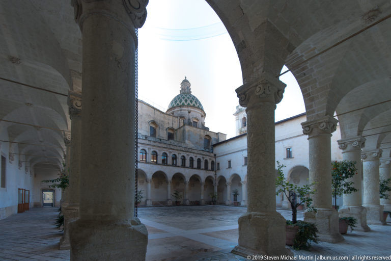 Stone cloister with rounded arcades and thick columns framing a courtyard, with a patterned tiled dome rising beyond.