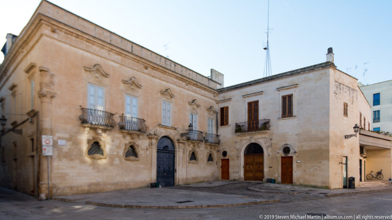 Corner limestone palazzo with iron balconies, carved window surrounds and multiple doorways facing an open paved space.
