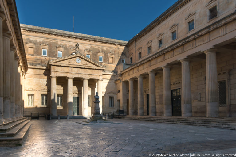 Large stone courtyard with pedimented portico, long classical colonnade and a small central monument in late-day light.