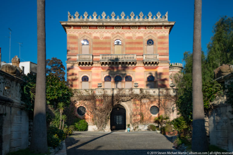 Polychrome historic villa with arched windows, balconies and decorative roof cresting behind a garden wall and tall palms.