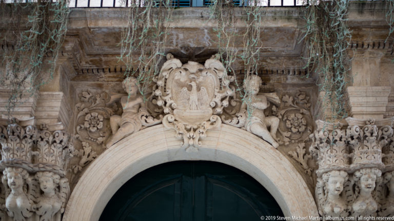 Ornate stone doorway with crowned cartouche, smaller shields, floral carving and trailing vegetation above a dark arched door.