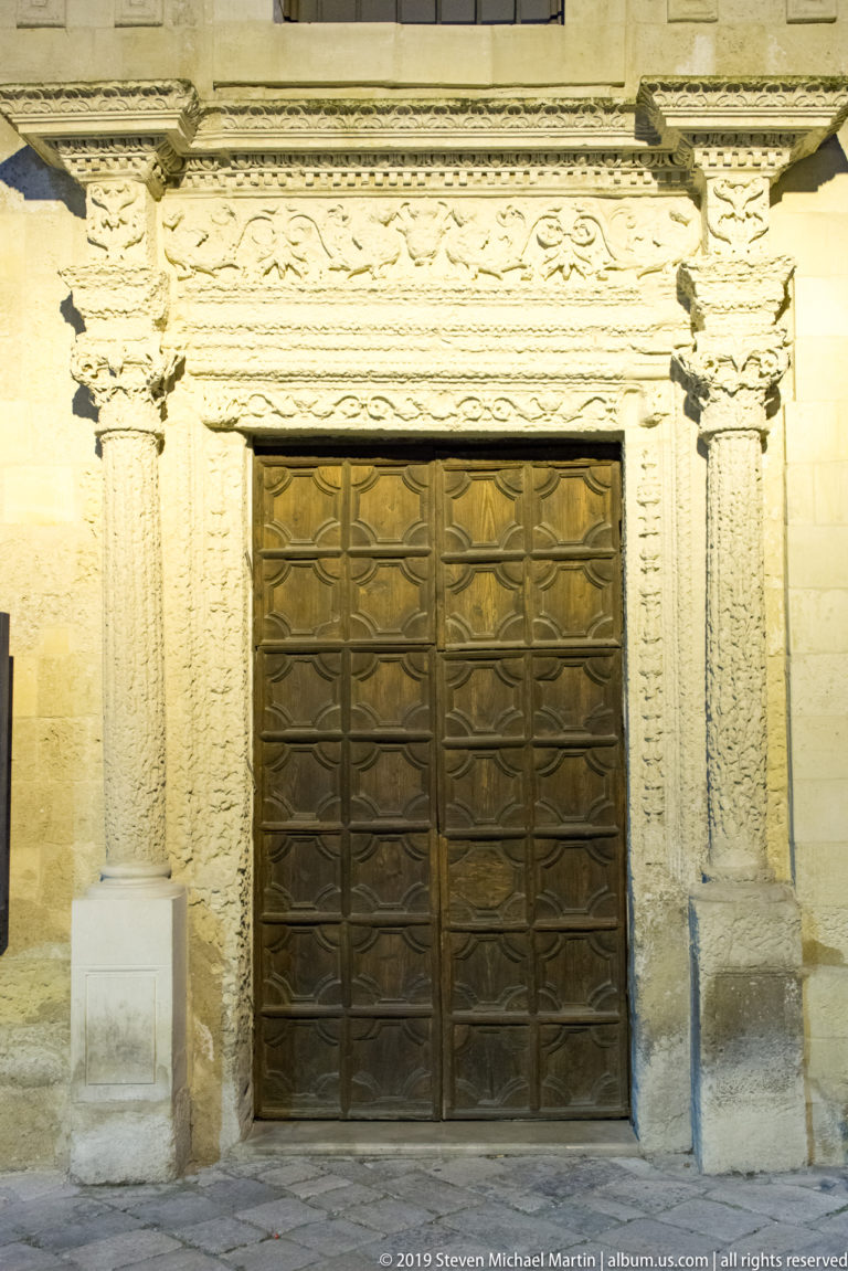 Dark paneled double doors set within a richly carved pale-stone portal decorated with floral and foliate relief.