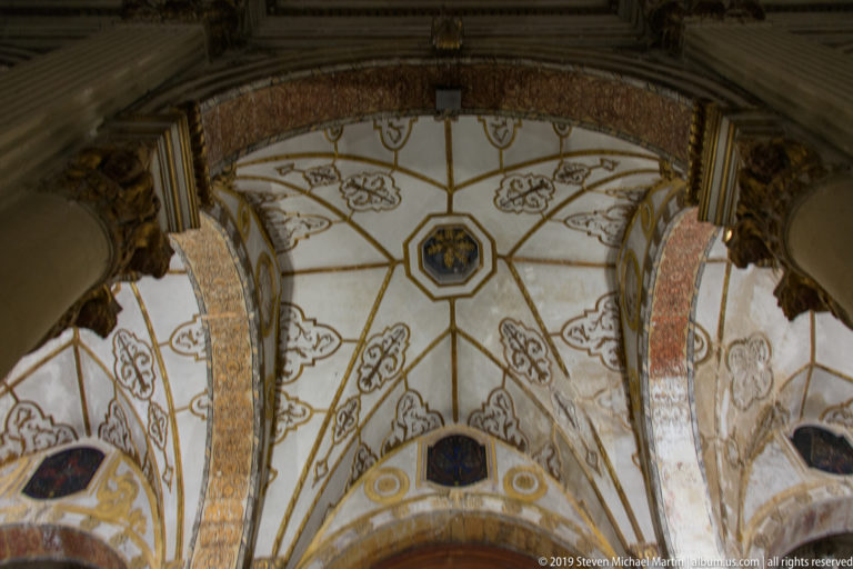 Upward view of a pale vaulted ceiling divided by gold ribs, with foliate motifs, small medallions, and decorated arches.