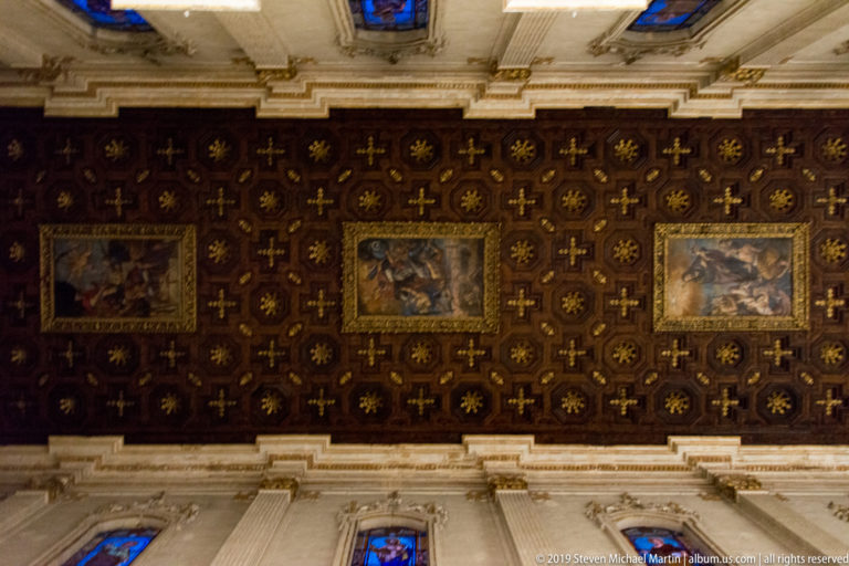 Dark wooden ceiling covered with gilded crosses and rosettes, with three rectangular religious paintings inset across the center.